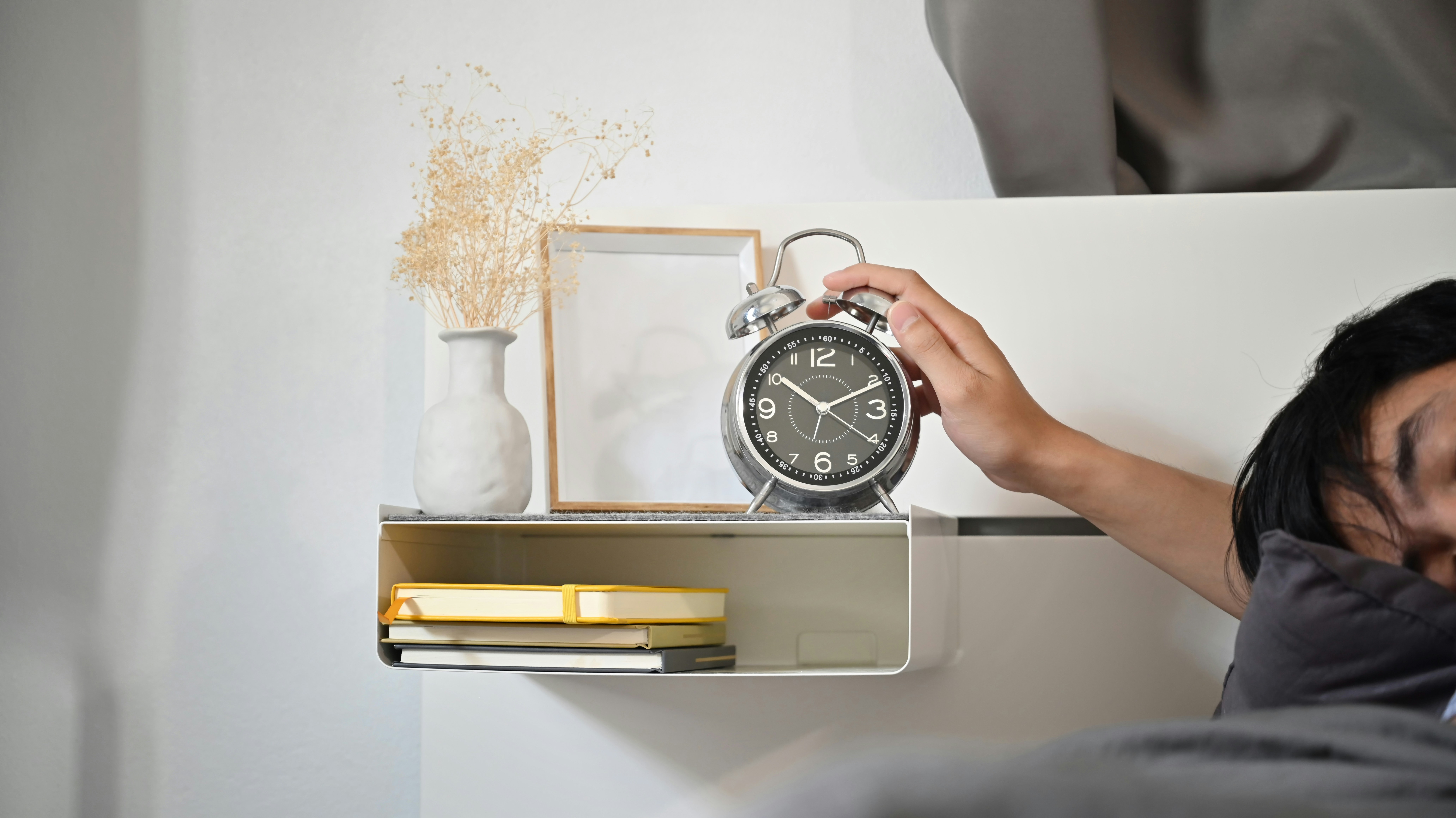 A person lying in bed reaching out to an alarm clock on a bedside shelf, the daily moment that sets sleep regularity